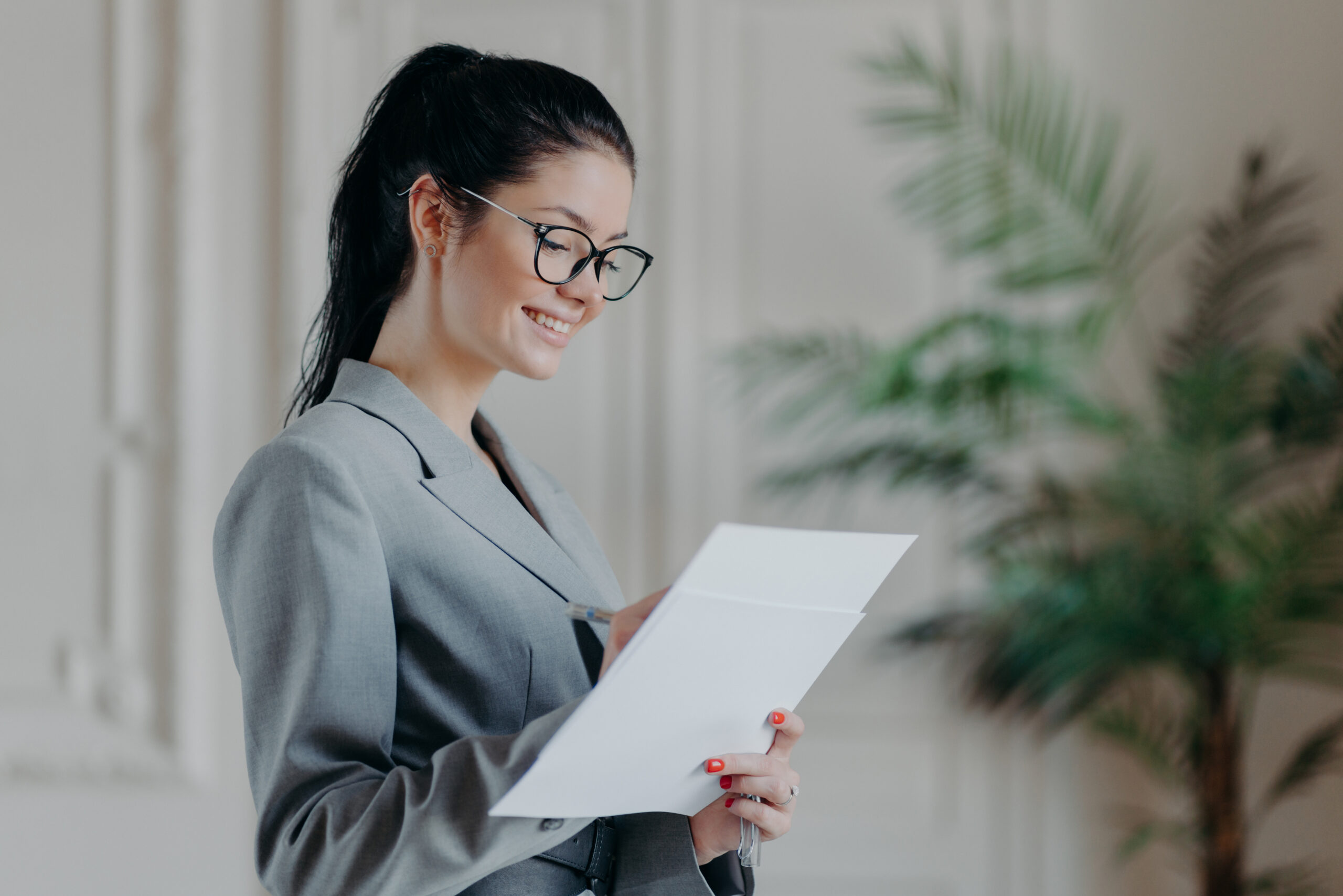 Glad female lawyer writes in corporate paper documents, wears optical glasses, formal clothes, stands in own cabinet, develops new strategy, has happy expression. People, career, employment concept